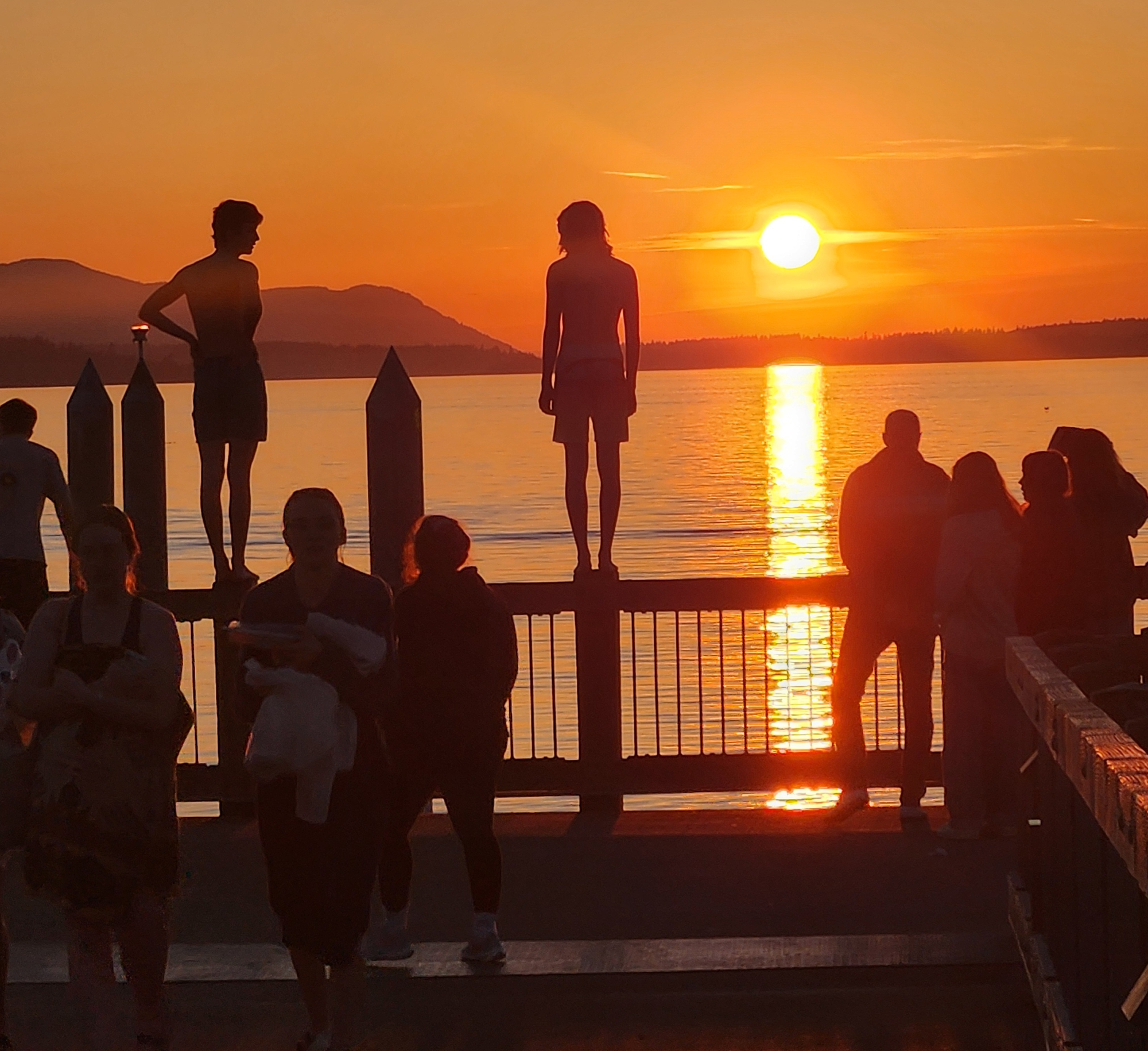 People stand on a railing with others nearby looking at the sunset over the waters of Bellingham Bay.