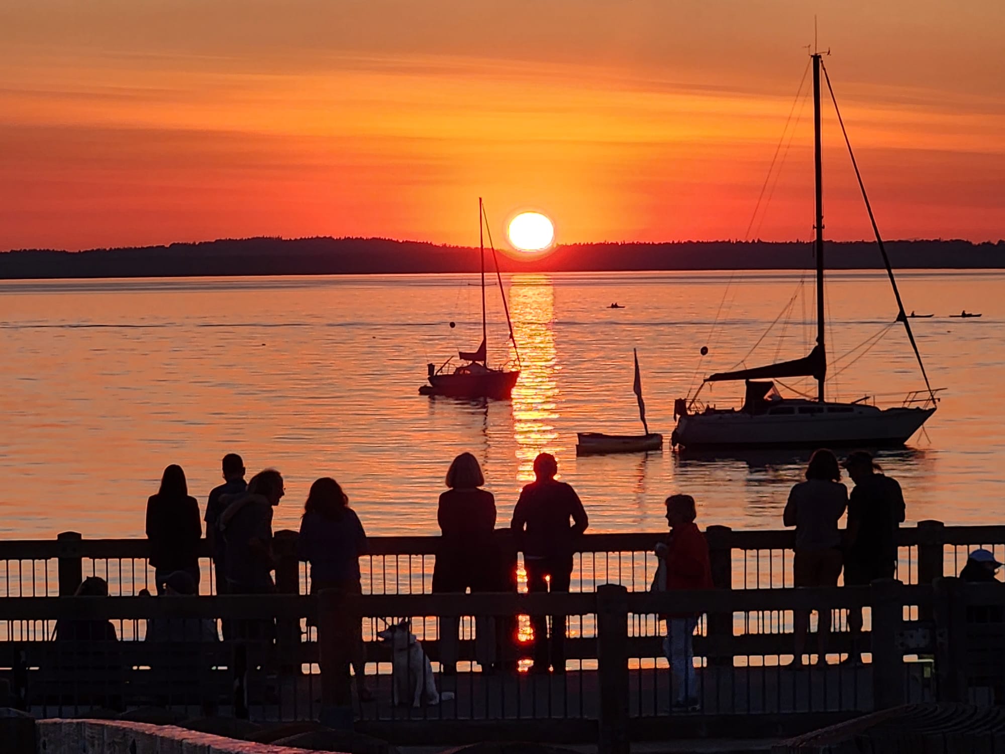 Silhouettes of people standing on a dock looking at boats on Bellingham Bay during sunset.