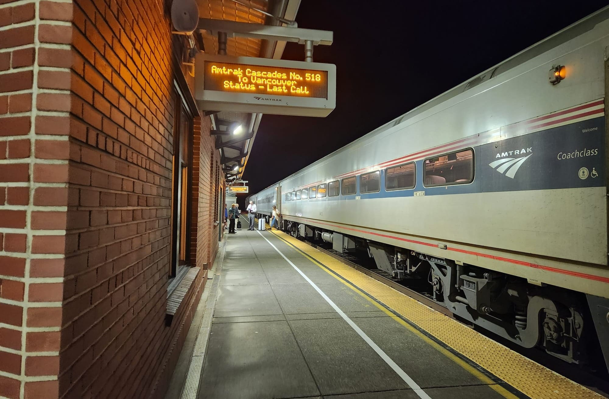 An Amtrak Cascades train at a train platform with a digital sign that reads "Amtrak Cascades No. 518 / To Vancouver / Status - Last Call"