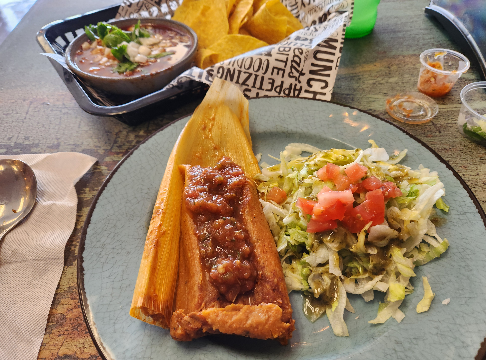 A pale blue plate with a traditional pork tamale covered in red chile with a bowl of pozole in the background.