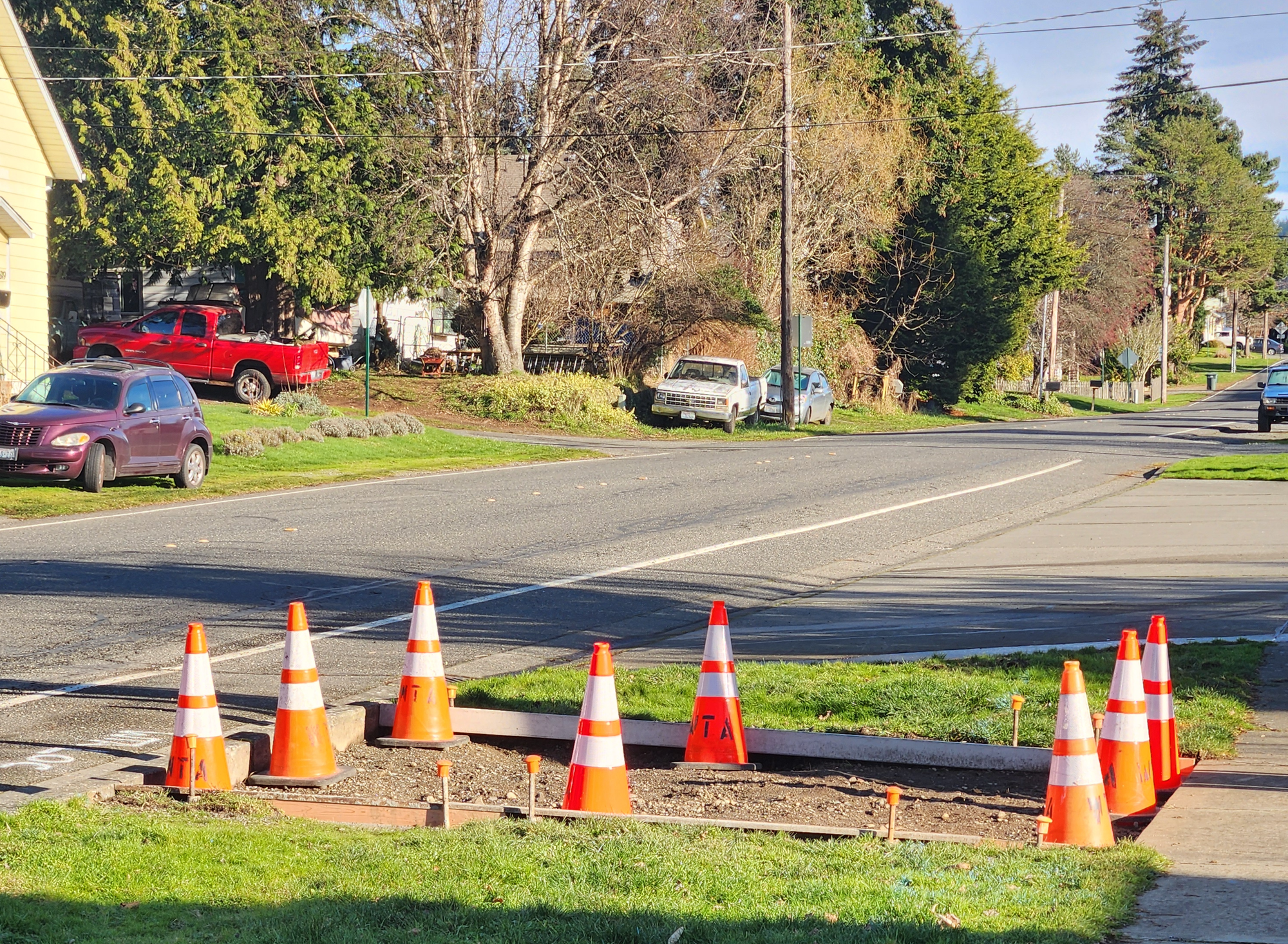 Orange construction cones mark off a section of sidewalk with a roadway in the background with cars parked on the grass. 